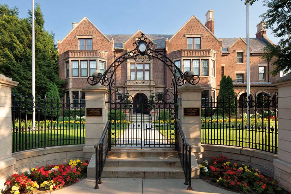 A view from the sidewalk in front of the Governor's Residence. Behind the fence is a large multi-story, symmetrical red brick mansion. Large windows adorn the front.
