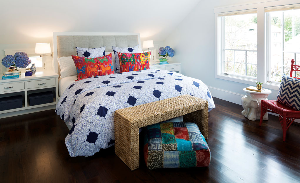 A master bedroom with sloping ceiling features a king size bed flanked by matching white nightstands. A wicker bench sits at the foot of the bed, and a red chair sits next to the large windows.