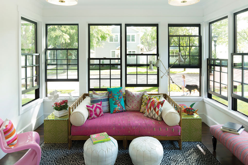 The front porch features large windows that look out onto the sidewalk and street. The inside is accented with a pink couch with colorful pillows, a pink chair and bench.