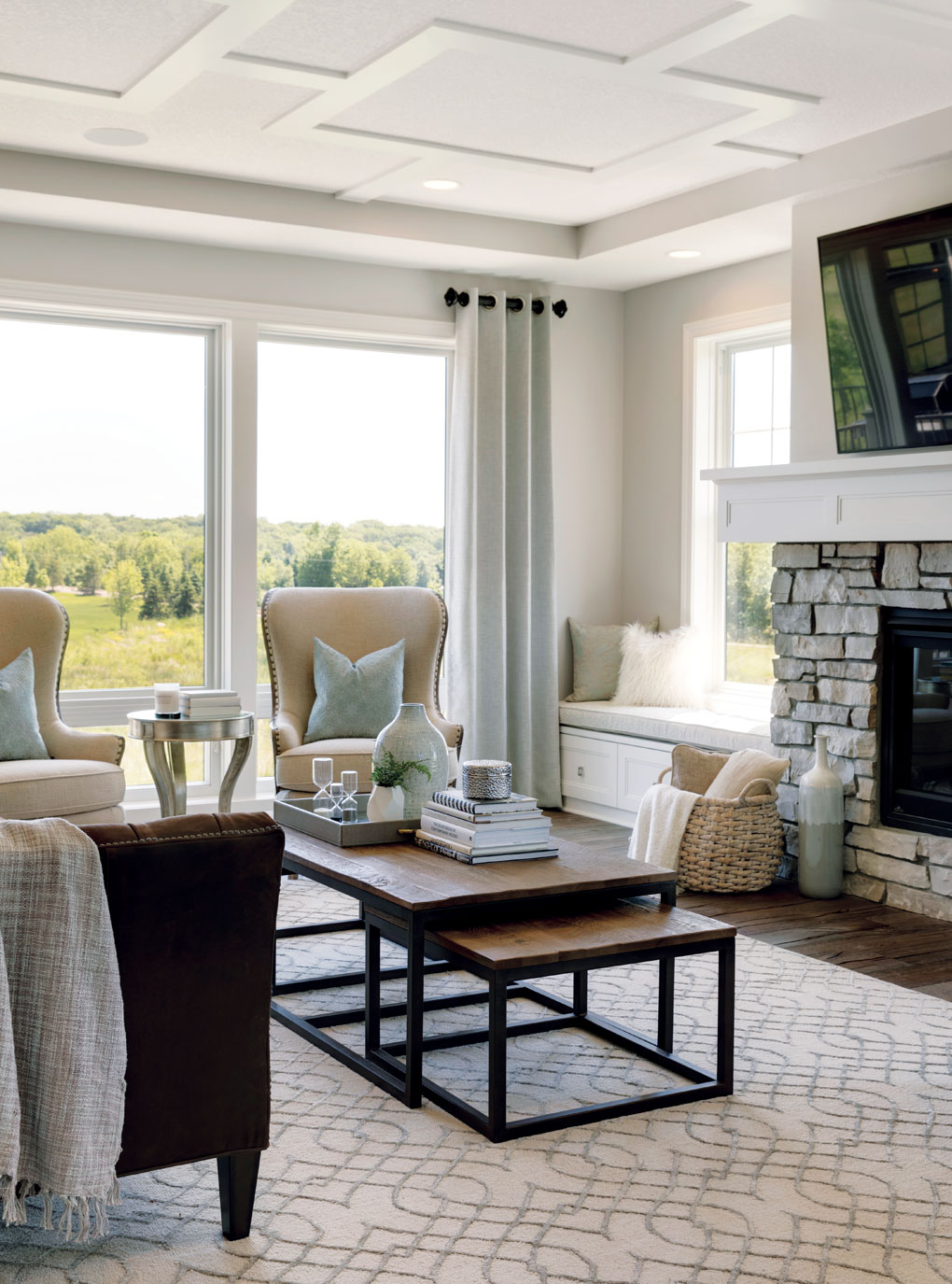 A great room with a coffered ceiling, floor to ceiling windows, and stone fireplace is a comfortable, welcoming space.