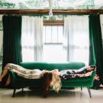 Jennifer Jorgensen and Kate Worum of She She lie on a green couch in a room covered in wallpaper featuring hand drawn chandeliers.